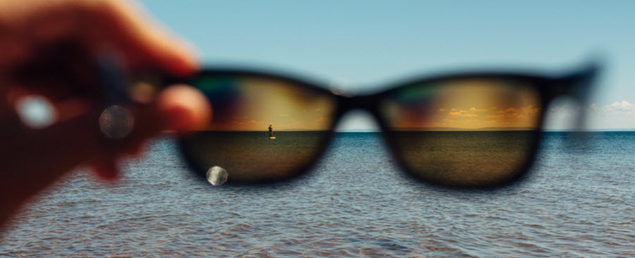 Sunglasses held up to the camera through which a person is seen paddle boarding in the distance.