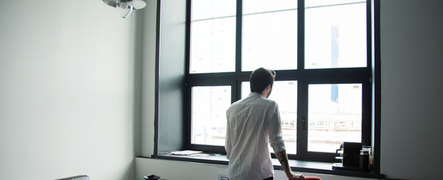 Man standing in his office looking outside a large window.