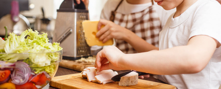 Un garçon tranche de la viande pendant qu'une fille râpe du fromage en arrière-plan.