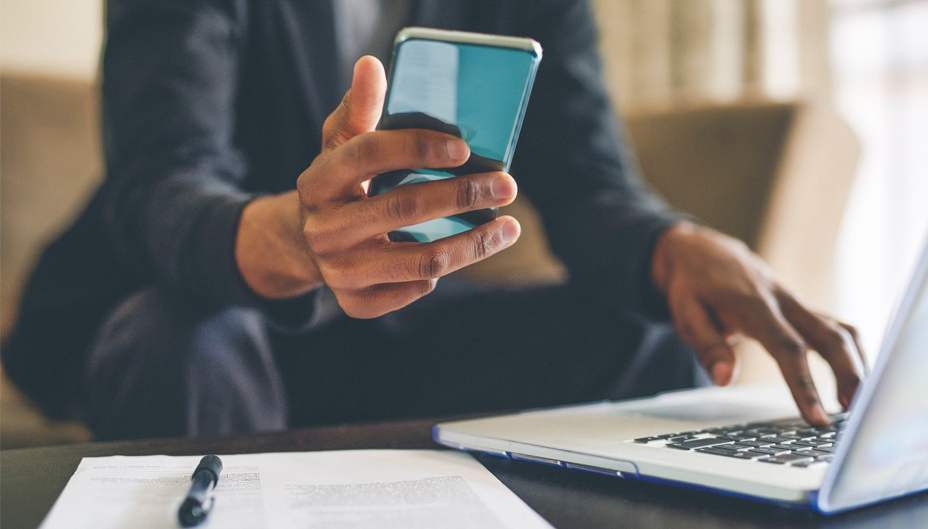 A close-up photo of someone working on a laptop with a cellphone in their other hand.