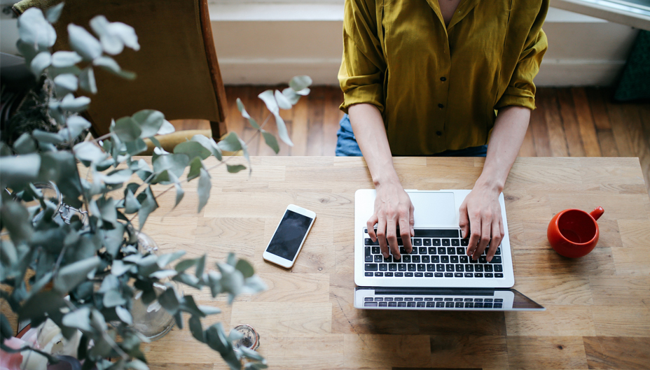 A woman working on a laptop, with a phone and cup of coffee nearby.