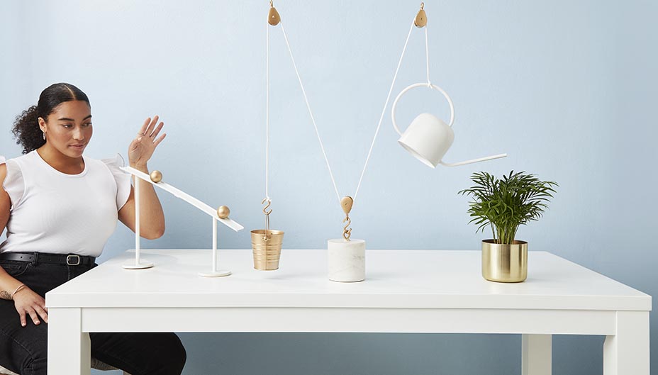 Woman pushing a ball through a Rube Goldberg machine to water a plant. 