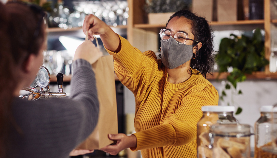 A masked retail worker hands over a shopping bag to a customer.