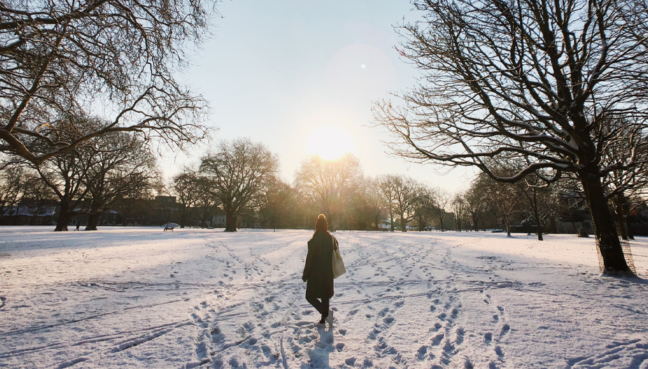 A woman walking through a snowy park during a sunny day.