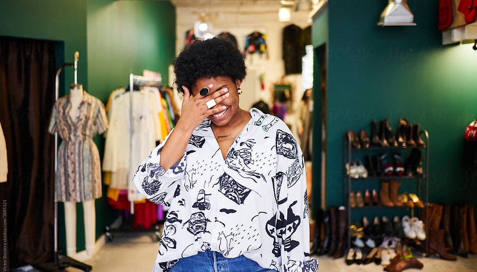 A Black businesswoman stands in a store with a hand held over her face, revealing only a smile.